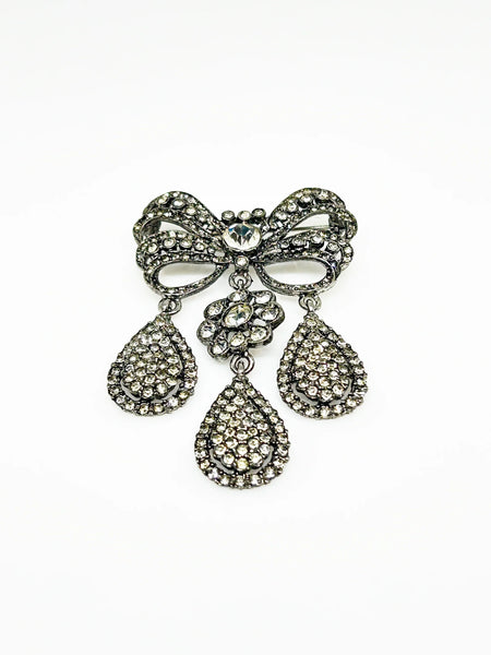 This exquisite piece of jewelry, crafted by Kenneth Jay Lane, is a shimmering testament to the exquisite beauty of the 18th century. It is dazzling, with crystal studded bows and gracefully dangling elaborate teardrops.The brooch is set in silver tone metal, its regal size measures 3.25" at its longest drop and 2" at its widest. The designer's signature is embossed on a raised oval at its back. Free Shipping