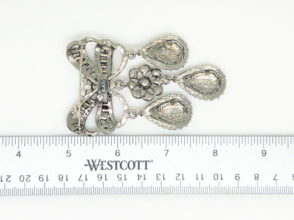 This exquisite piece of jewelry, crafted by Kenneth Jay Lane, is a shimmering testament to the exquisite beauty of the 18th century. It is dazzling, with crystal studded bows and gracefully dangling elaborate teardrops.The brooch is set in silver tone metal, its regal size measures 3.25" at its longest drop and 2" at its widest. The designer's signature is embossed on a raised oval at its back. Free Shipping