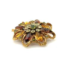 A 1950s Austrian costume jewelry flower pin featuring tiered, multi-colored faceted crystals in warm amber, yellow, and brown tones. The center cluster is arranged with small, round olivine green rhinestones, set on a gold-tone filigree metal backing against a plain white background.
