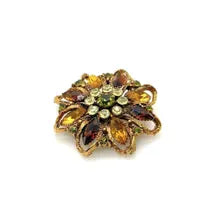 A 1950s Austrian costume jewelry flower pin featuring tiered, multi-colored faceted crystals in warm amber, yellow, and brown tones. The center cluster is arranged with small, round olivine green rhinestones, set on a gold-tone filigree metal backing against a plain white background.
