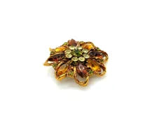 A 1950s Austrian costume jewelry flower pin featuring tiered, multi-colored faceted crystals in warm amber, yellow, and brown tones. The center cluster is arranged with small, round olivine green rhinestones, set on a gold-tone filigree metal backing against a plain white background.
