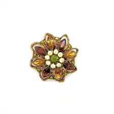 A 1950s Austrian costume jewelry flower pin featuring tiered, multi-colored faceted crystals in warm amber, yellow, and brown tones. The center cluster is arranged with small, round olivine green rhinestones, set on a gold-tone filigree metal backing against a plain white background.