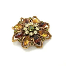 A 1950s Austrian costume jewelry flower pin featuring tiered, multi-colored faceted crystals in warm amber, yellow, and brown tones. The center cluster is arranged with small, round olivine green rhinestones, set on a gold-tone filigree metal backing against a plain white background.