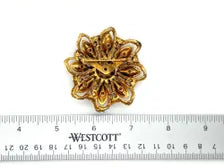 A small gold-toned flower-shaped brooch or pin placed above a white measuring ruler for scale, with the word WESTCOTT visible on the ruler.