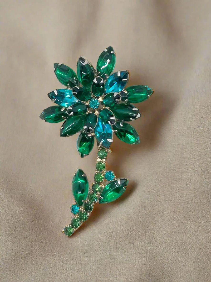 This breathtaking floral brooch embodies the essence of Juliana style with its vibrant gold tone and dazzling rhinestones. The combination of green and teal glass stones creates a mesmerizing color scheme, and its size of 3 x 1.75 x 0.625 inches is perfect for making a statement. 