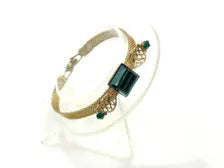 Vintage gold-tone mesh bracelet featuring a large, translucent emerald-cut green glass crystal centerpiece with delicate filigree side accents on a white display stand