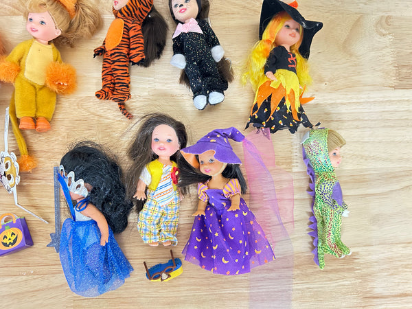 A collection of vintage Halloween dolls and costume accessories arranged on a light wood floor. The dolls are dressed in various costumes including a yellow monster, a tiger, a black sparkly dress, a witch with yellow hair, a blue dress, a purple moonlit dress, and a green dinosaur.