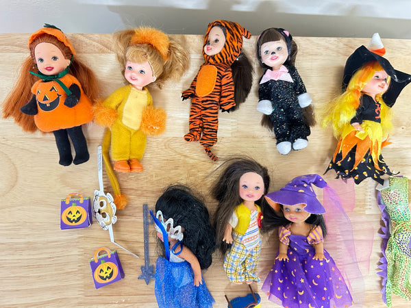 A collection of vintage Halloween-themed dolls and accessories arranged on a light wood surface. The dolls are dressed in various costumes including pumpkin, tiger, black cat, witch, and purple wizard outfits, with small jack-o'-lantern bags and a tiara.