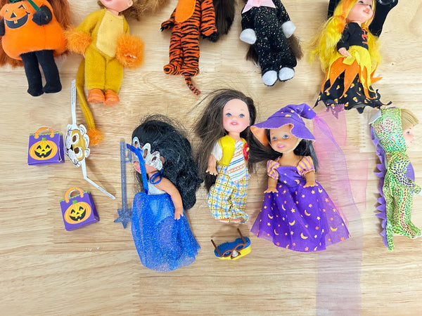 A collection of vintage Halloween-themed dolls and accessories arranged on a light-colored wooden floor. The dolls are dressed in various costumes including orange, yellow, black, purple, and green, with small pumpkin bags and a toy sword.