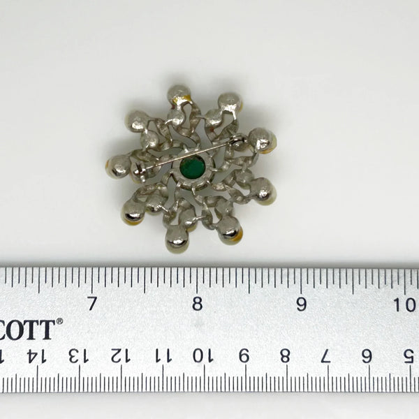 This exquisite brooch showcases the artistry of a master costume jeweler. It exudes a regal aura, as if it were plucked from a royal vault. Enhanced by a green round cabochon and sparkling swirls, accented with cream faux pearls and crafted in silver tone metal, this piece measures 1 3/4" in width and is in impeccable condition.