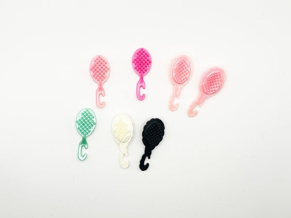 A top-down view of seven vintage miniature doll hairbrushes in various shades of pink, mint green, white, and black, neatly arranged on a solid white background. The plastic toy brushes feature a distinct molded C-shaped hook handle designed to hang from a doll's wrist or vanity set