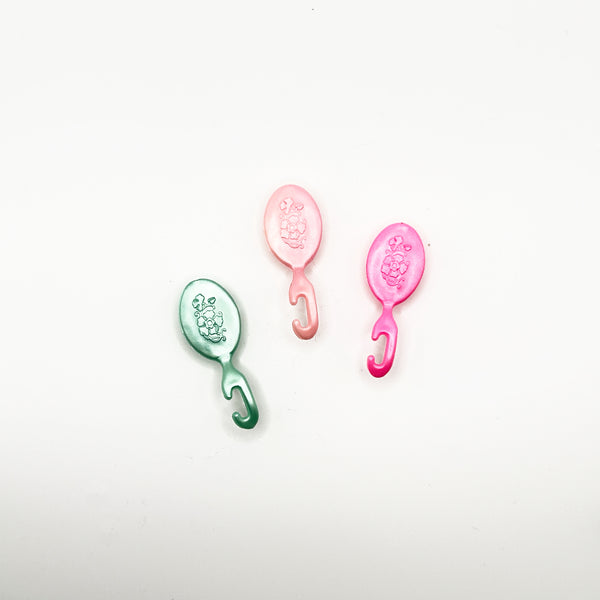 A collection of three miniature plastic doll styling accessories from the 1980 Mattel Western Barbie line. The lot includes a metallic teal green brush, a pale pastel pink brush, and a vibrant hot pink brush. Each item is molded with an intricate, traditional floral pattern on the back and ends in a curved hook-style clip handle meant to clip onto a doll's hand