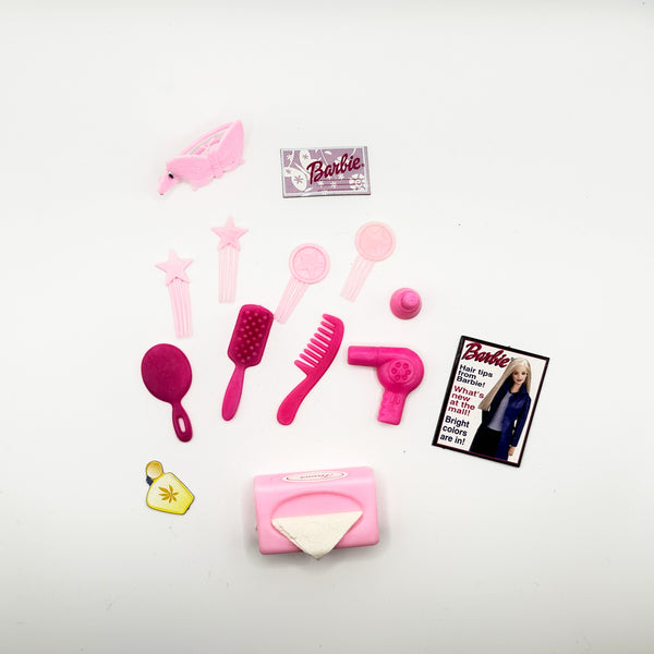 A mixed lot of vintage Barbie doll hair styling and vanity accessories arranged on a white background, featuring various pink brushes, combs, a miniature toy hairdryer, accessory bottles, a small paper Barbie styling booklet, and a black hook brush