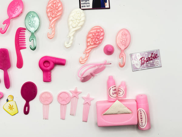 A Barbie-themed miniature toy hair salon set arranged on a white background, including pink hairbrushes, combs, a hair dryer, hair clips, a small pink bottle labeled shampoo, a pink towel, and a small Barbie card.