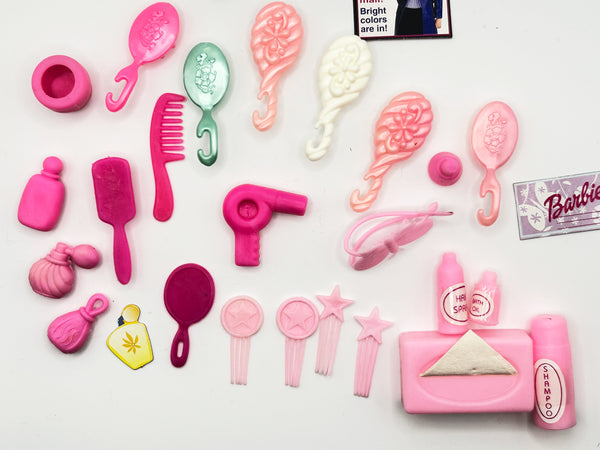 A mixed lot of vintage Barbie doll hair styling and vanity accessories arranged on a white background, featuring various pink brushes, combs, a miniature toy hairdryer, accessory bottles, a small paper Barbie styling booklet, and a black hook brush