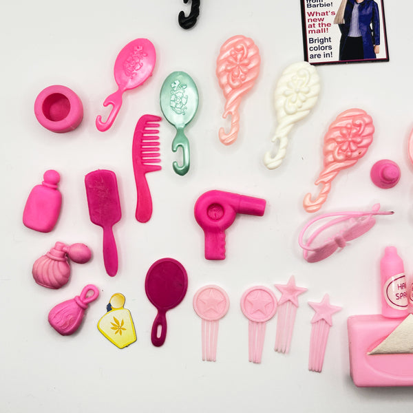 A mixed lot of vintage Barbie doll hair styling and vanity accessories arranged on a white background, featuring various pink brushes, combs, a miniature toy hairdryer, accessory bottles, a small paper Barbie styling booklet, and a black hook brush