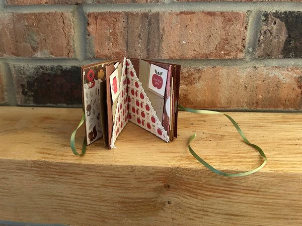 Handmade mini autumn scrapbook album with an orange and green striped patterned cover, featuring a cartoon fox illustration, a die-cut red maple leaf, a gold heart, and the text "HELLO FRIEND." A green satin ribbon closure extends from the side of the book, which sits on a wooden shelf against a brick background.
