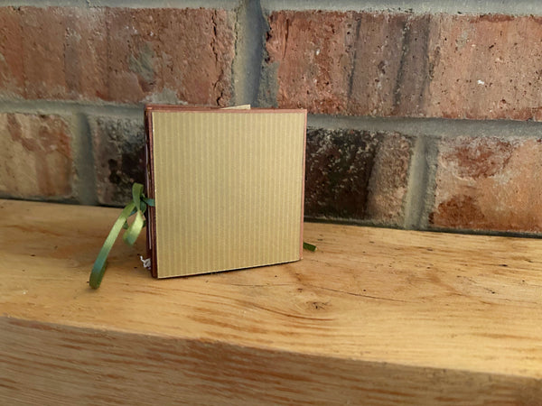 Handmade mini autumn scrapbook album with an orange and green striped patterned cover, featuring a cartoon fox illustration, a die-cut red maple leaf, a gold heart, and the text "HELLO FRIEND." A green satin ribbon closure extends from the side of the book, which sits on a wooden shelf against a brick background.
