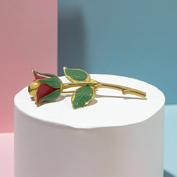 Gold rose brooch with red and green details on a white pedestal against a pink and blue background