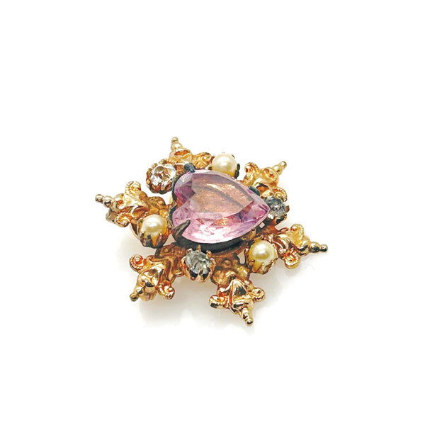 A gold-toned brooch with a central heart-shaped, light purple gemstone. The brooch is surrounded by ornate gold filigree, small pearls, and clear crystals, creating a vintage, elegant design.