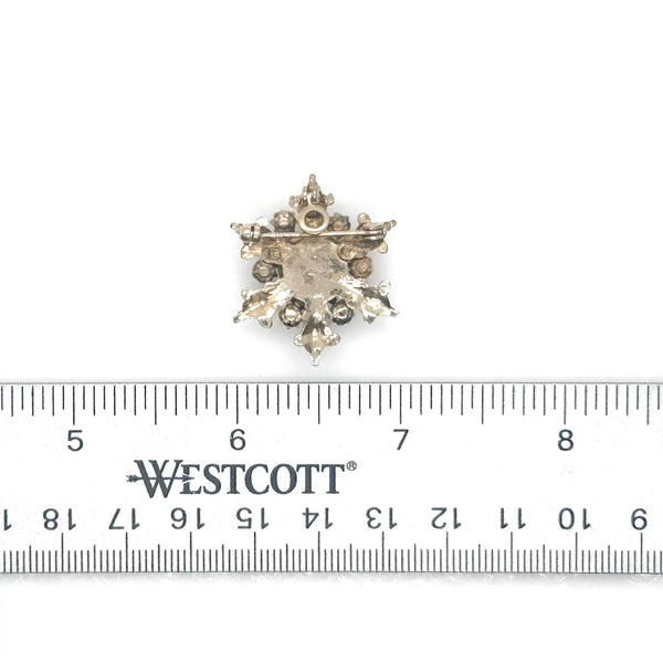 A gold-toned brooch with a central heart-shaped, light purple gemstone. The brooch is surrounded by ornate gold filigree, small pearls, and clear crystals, creating a vintage, elegant design.