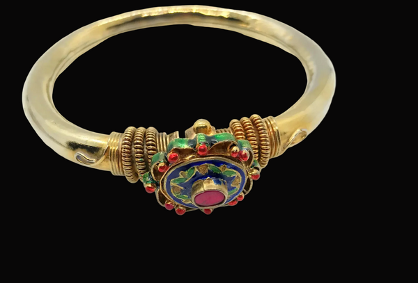 A gold bangle bracelet featuring a central decorative clasp with blue enamel, red stones, and green accents, shown against a dark gray background.