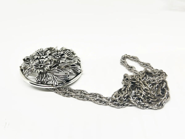 This striking, rounded, and very detailed brooch pendant makes a beautiful necklace. It measures 2" across and comes on a 24" chain with a secure fold-over clasp. The pendant features a strong, eye-catching frame that showcases a delicate floral motif, complete with blooms and leaves