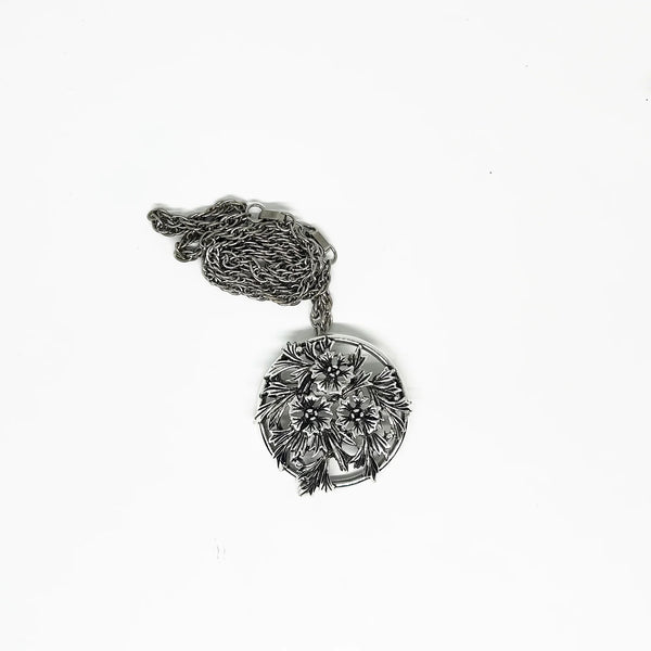 This striking, rounded, and very detailed brooch pendant makes a beautiful necklace. It measures 2" across and comes on a 24" chain with a secure fold-over clasp. The pendant features a strong, eye-catching frame that showcases a delicate floral motif, complete with blooms and leaves