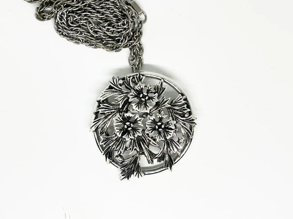 This striking, rounded, and very detailed brooch pendant makes a beautiful necklace. It measures 2" across and comes on a 24" chain with a secure fold-over clasp. The pendant features a strong, eye-catching frame that showcases a delicate floral motif, complete with blooms and leaves