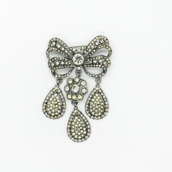 This exquisite piece of jewelry, crafted by Kenneth Jay Lane, is a shimmering testament to the exquisite beauty of the 18th century. It is dazzling, with crystal studded bows and gracefully dangling elaborate teardrops.The brooch is set in silver tone metal, its regal size measures 3.25" at its longest drop and 2" at its widest. The designer's signature is embossed on a raised oval at its back. Free Shipping