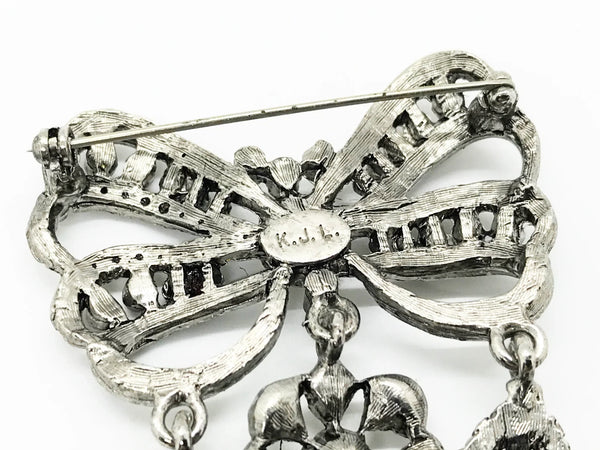 This exquisite piece of jewelry, crafted by Kenneth Jay Lane, is a shimmering testament to the exquisite beauty of the 18th century. It is dazzling, with crystal studded bows and gracefully dangling elaborate teardrops.The brooch is set in silver tone metal, its regal size measures 3.25" at its longest drop and 2" at its widest. The designer's signature is embossed on a raised oval at its back. Free Shipping