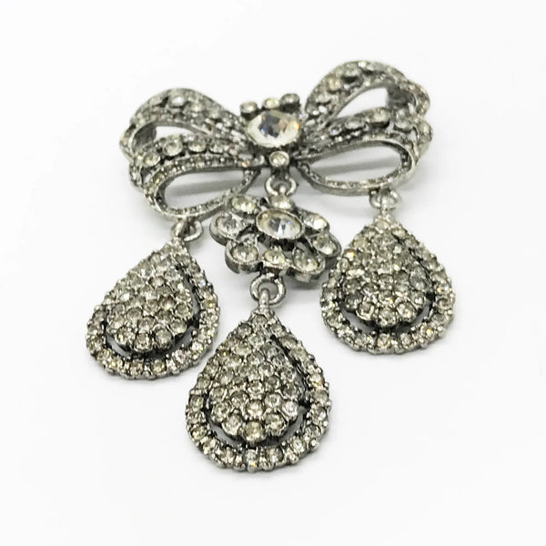 This exquisite piece of jewelry, crafted by Kenneth Jay Lane, is a shimmering testament to the exquisite beauty of the 18th century. It is dazzling, with crystal studded bows and gracefully dangling elaborate teardrops.The brooch is set in silver tone metal, its regal size measures 3.25" at its longest drop and 2" at its widest. The designer's signature is embossed on a raised oval at its back. Free Shipping