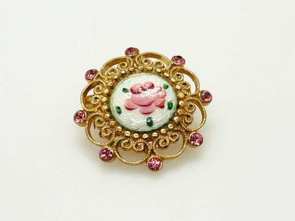 Take a look at this vintage costume jewelry brooch featuring a hand-painted pink rose on a white enamel or ceramic base, set within an ornate gold-tone filigree frame accented with pink rhinestones. The center showcases a pink rose, with green dots accenting the perimeter. The setting and metalwork around the outside are particularly lovely. It measures 1" across.