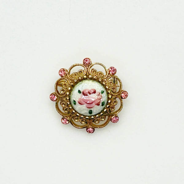 Take a look at this vintage costume jewelry brooch featuring a hand-painted pink rose on a white enamel or ceramic base, set within an ornate gold-tone filigree frame accented with pink rhinestones. The center showcases a pink rose, with green dots accenting the perimeter. The setting and metalwork around the outside are particularly lovely. It measures 1" across.