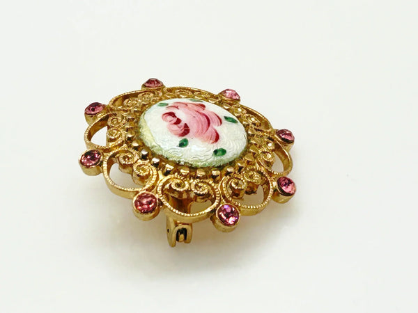 Take a look at this vintage costume jewelry brooch featuring a hand-painted pink rose on a white enamel or ceramic base, set within an ornate gold-tone filigree frame accented with pink rhinestones. The center showcases a pink rose, with green dots accenting the perimeter. The setting and metalwork around the outside are particularly lovely. It measures 1" across.