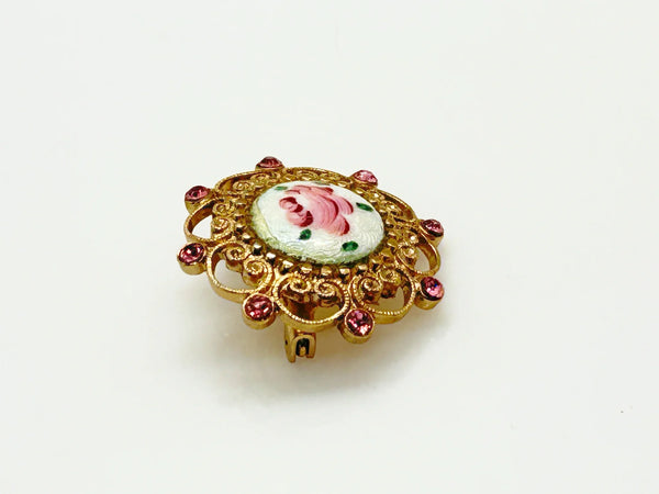 Take a look at this vintage costume jewelry brooch featuring a hand-painted pink rose on a white enamel or ceramic base, set within an ornate gold-tone filigree frame accented with pink rhinestones. The center showcases a pink rose, with green dots accenting the perimeter. The setting and metalwork around the outside are particularly lovely. It measures 1" across.