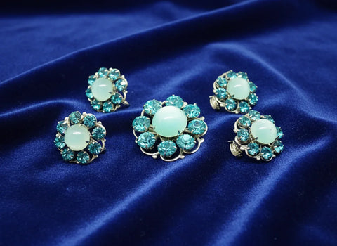 This is a hard-to-find set featuring a brooch, a pair of scatter pins, and screw-back earrings. The design showcases round, frosted light turquoise cabochons. Each features a center moonglow stone surrounded by bright, faceted, slightly deeper blue stones, all prong-set. 