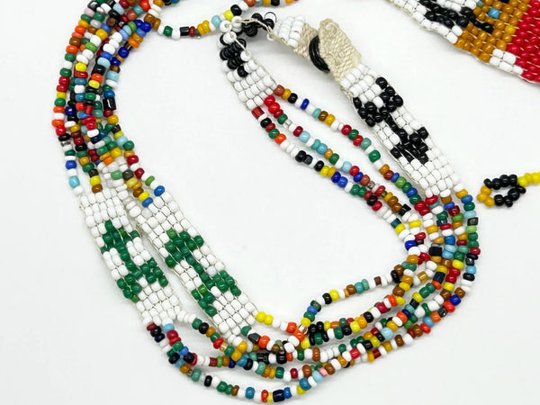 This striking Native American beaded necklace features a beautifully detailed figure adorned with two feathers in flowing black hair, dressed in a brown tunic and red skirt with black shoes. The design showcases a white upper background that transitions to a vibrant turquoise-teal green below, creating visual depth and interest. 
