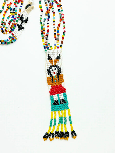 This striking Native American beaded necklace features a beautifully detailed figure adorned with two feathers in flowing black hair, dressed in a brown tunic and red skirt with black shoes. The design showcases a white upper background that transitions to a vibrant turquoise-teal green below, creating visual depth and interest. 
