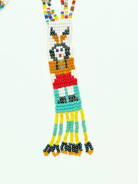 This striking Native American beaded necklace features a beautifully detailed figure adorned with two feathers in flowing black hair, dressed in a brown tunic and red skirt with black shoes. The design showcases a white upper background that transitions to a vibrant turquoise-teal green below, creating visual depth and interest. 