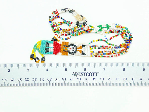 This striking Native American beaded necklace features a beautifully detailed figure adorned with two feathers in flowing black hair, dressed in a brown tunic and red skirt with black shoes. The design showcases a white upper background that transitions to a vibrant turquoise-teal green below, creating visual depth and interest. 