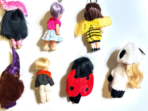 Six small doll figures or doll accessories arranged on a plain white background, each with different hair, clothing, and costume details visible from the back.