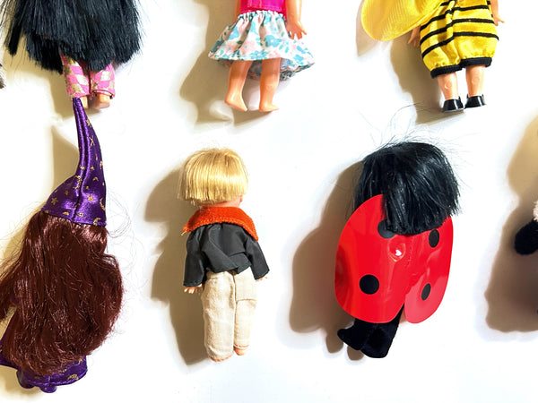 A group of vintage dolls and doll accessories arranged on a plain white background, including a child doll in a black and orange outfit, a bee-themed outfit, a wizard hat with long brown hair, and a red ladybug costume.