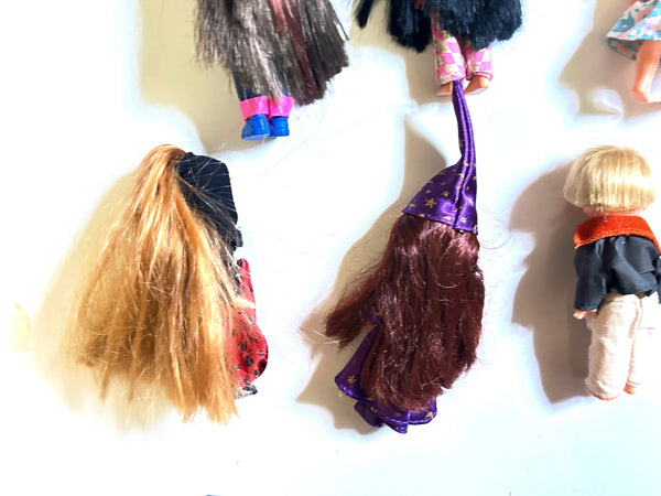 Several doll heads and hair pieces laid out on a white surface, including one with long blonde hair, one with dark brown hair and a purple witch hat, and another partially visible at the top.