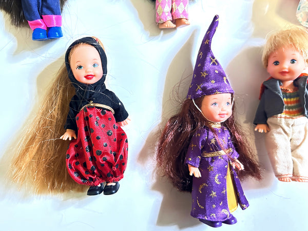 Two small dolls dressed in costumes posed on a white surface. One doll wears a black outfit with a red patterned skirt, and the other wears a purple wizard costume with gold stars.