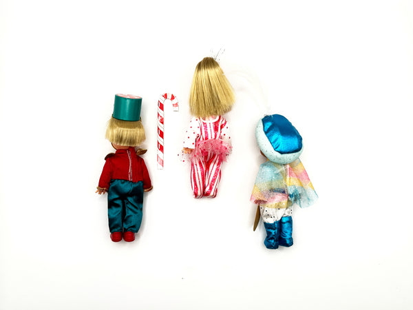 Back angle of three 4.5-inch Kelly Club miniature figures. On the left, Tommy as Major Mint shows the rear construction of his red and teal toy soldier uniform. The center Kelly doll displays the reverse side of her pink-and-white striped dress and a clear view of her blonde hair bundle. On the right, the African American edition Tommy doll shows the back of his white-furred winter coat hood and blue uniform pants