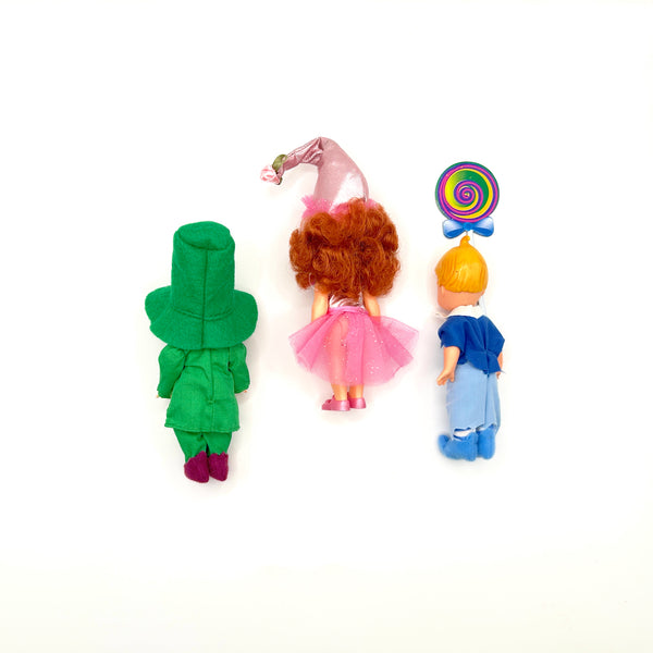 Three loose vintage 1999 Mattel Barbie Kelly Club mini dolls from The Wizard of Oz line arranged on a white background: Tommy as Mayor Munchkin in a green felt top hat and jacket on the left, Kelly as Lullaby Munchkin in a pink conical hat and tutu in the center, and Tommy as Lollipop Munchkin holding a colorful swirl lollipop on the right