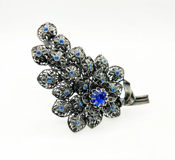This exquisite vintage pewter tone blue rhinestone floral brooch boasts dimensions of 4" by 2.25" and features three stems and a partial bow. The tiered design showcases stunning, prong-set blue rhinestones in the form of a large blossom, complemented by bezel-set stones in each smaller blossom.