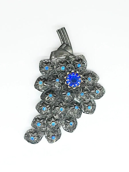 This exquisite vintage pewter tone blue rhinestone floral brooch boasts dimensions of 4" by 2.25" and features three stems and a partial bow. The tiered design showcases stunning, prong-set blue rhinestones in the form of a large blossom, complemented by bezel-set stones in each smaller blossom.