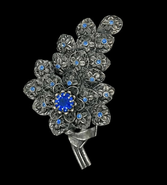 This exquisite vintage pewter tone blue rhinestone floral brooch boasts dimensions of 4" by 2.25" and features three stems and a partial bow. The tiered design showcases stunning, prong-set blue rhinestones in the form of a large blossom, complemented by bezel-set stones in each smaller blossom.